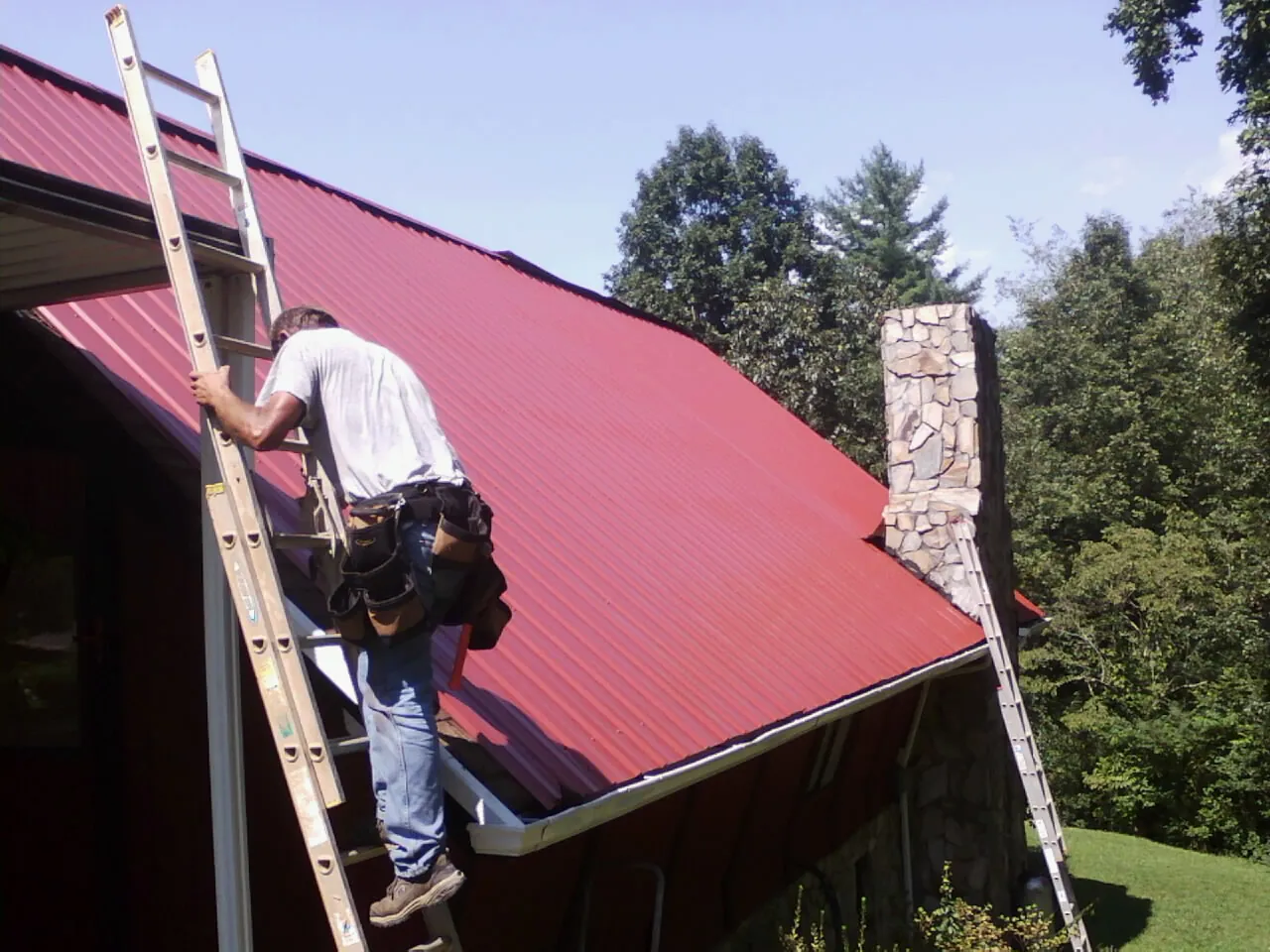 Roofer on a ladder beside a red metal roof and stone chimney — Soffit & Fascia Repair by Roofing Brothers in Banks